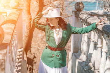 Woman walks around the city, lifestyle. Happy woman in a green jacket, white skirt and hat is sitting on a white fence with balusters overlooking the sea bay and the city.