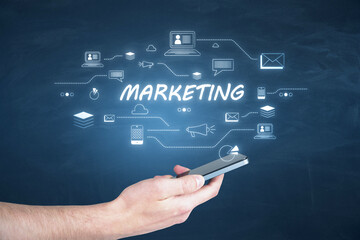 Hand holding a smartphone with MARKETING text and related icons on a virtual screen. Digital strategy concept