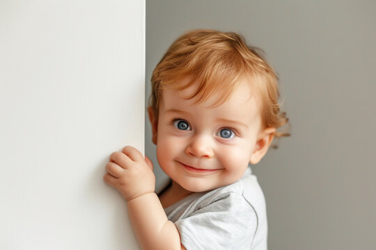 A Small Child, 1 Year Old, Stands Holding Onto The Wall And Looks Out From Behind The White Wall. Hide And Seek Games. Happy Childhood. The Child Looks At The Camera, Smiles.