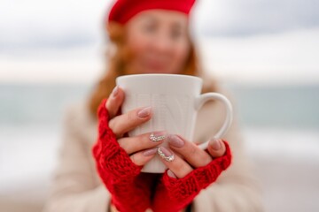 Woman beach in red beret scarf and mitts holding a cup of tea in his hands. Depicting beach relaxation and cozy attire. Walks by the sea