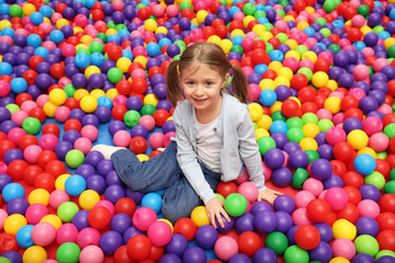 Happy little girl sitting on colorful balls in ball pit
