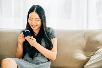 Young Asian woman seated on a cozy couch at home happily texts on her smartphone. Engaged in messaging online shopping and chatting. Embracing modern technology for communication.