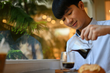 Cropped image of Asian male pouring freshly brewed coffee from pitcher to glass in the cafe