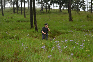 Wide angle view of young Asian hiker with backpack taking photos of natural scenery in the middle of green grass of a lush forest, Solo travel concept