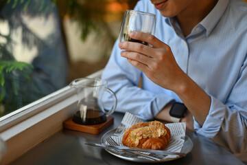 Cropped image of a Young man enjoying a black coffee and breakfast with croissant sitting on the table beside the window