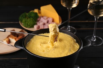 Dipping pieces of ham and bread into fondue pot with melted cheese on black wooden table, closeup