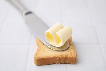 Tasty butter curl, knife and piece of dry bread on white tiled table, closeup