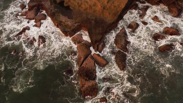 Top static view of the waves of the ocean crashing against stones coastline. The sea surf breaking on rocks and turning into a seething white foam. Water swirls around the shore