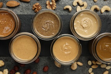 Tasty nut butters in jars and raw nuts on gray table, flat lay