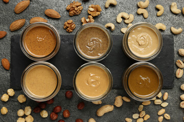 Tasty nut butters in jars and raw nuts on gray table, flat lay