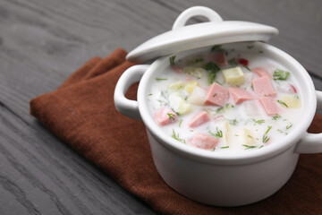 Delicious cold summer soup (okroshka) with boiled sausage in pot on grey wooden table, closeup....