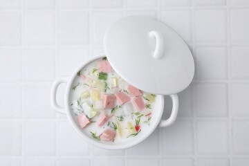 Delicious cold summer soup (okroshka) with boiled sausage in pot on white tiled table, top view