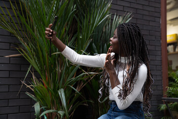 Portrait of beautiful young African American woman showing long black hair braided hairstyle and taking selfie on mobile phone while feeling happy and smiling in the city outdoor