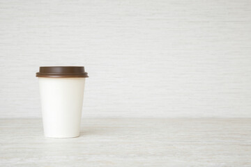 New white disposable paper cup with dark brown cap on stone table top. Takeaway coffee or other drink. Closeup. Empty place for text on light beige wallpaper background. Front view.