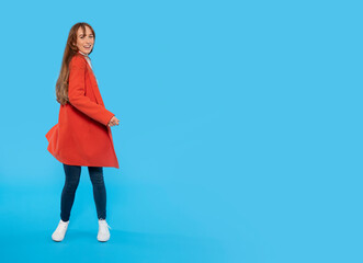 Joyful cheerful smiling attractive young redhead woman in jeans and orange coat standing, closing eyes isolated on a blue background studio portrait