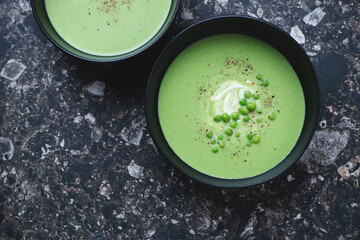 Bowls with green pea cream-soup on a dark-brown granite background, horizontal shot with space, flat lay