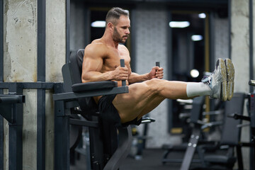 Handsome sportsman with hairstyle pulling up legs, working out indoors. Full body of muscular male athlete in shorts doing pull ups on training apparatus in gym. Concept of sport, workout.