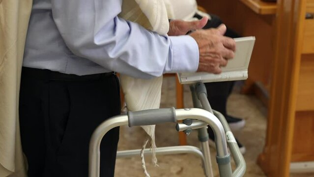 An elderly Jewish man is using a walker to pray at the Tora book and wearing a Talit.