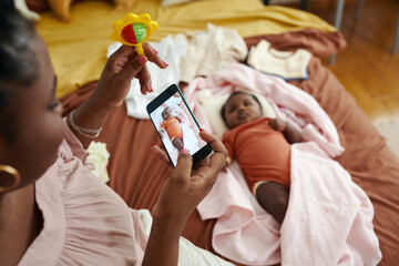 Mother shaking rattle to attract attention of baby and take photo for social media - obrazy, fototapety, plakaty