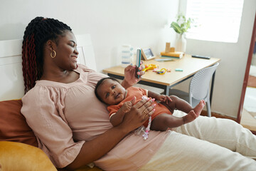 Smiling Black woman resting on bed with little baby and texting friends or checking social media