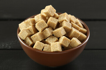 Brown sugar cubes in bowl on black wooden table, closeup