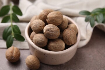 Whole nutmegs in bowl and green branches on brown table, closeup