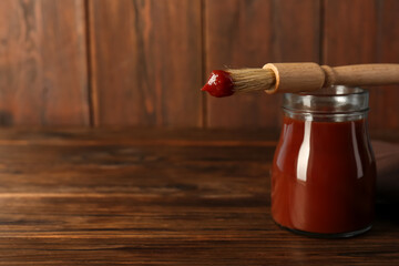 Tasty barbecue sauce in glass jar and brush on wooden table, closeup. Space for text
