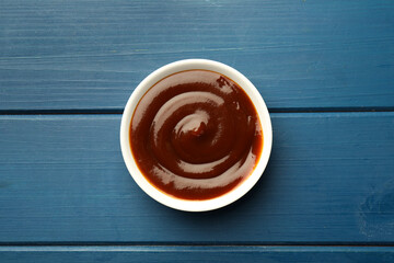 Tasty barbeque sauce in bowl on blue wooden table, top view