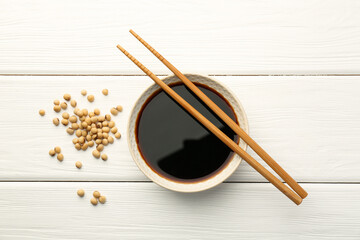 Tasty soy sauce in bowl, chopsticks and soybeans on white wooden table, top view