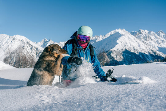 Female snowboarder playing with dog on ski resort, winter sport outdoor, sunny day in mountains