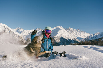 Female snowboarder playing with dog on ski resort, winter sport outdoor, sunny day in mountains