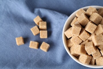 Brown sugar cubes in bowl on blue tablecloth, top view. Space for text