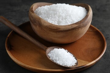 Fototapeta na wymiar Natural salt in wooden bowl and spoon on dark grey table, closeup
