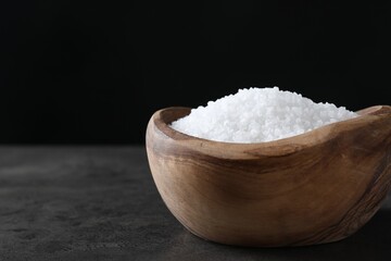 Natural salt in wooden bowl on dark grey table, closeup. Space for text