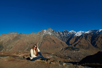 A gorgeous woman framed by tall, snow-topped brown mountains, looking down at a picturesque village...