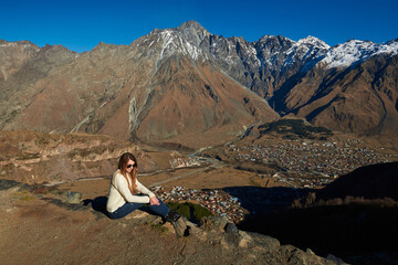  An attractive tourist admiring the breathtaking view of high brown mountains draped in snow from afar, with a charming small town visible below.