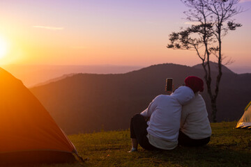 Couple is sitting by the tent during overnight camping and taking selfie while looking at the beautiful scenic sunset over the mountain for outdoor adventure vacation travel