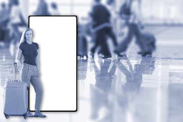 Young smiling woman is standing near a suitcase in an airport terminal. Blurred figures of walking passengers are in the background. Travel concept.