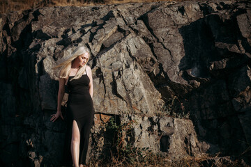 A slender, positive, sexy aged woman with long blond hair in a black long stylish dress stands near the picturesque rocks on a bright sunny summer day.