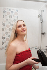 Nice blonde woman dries her hair with a hair dryer, in the bathroom. Creative image on the topic of health, hygiene, hair care.