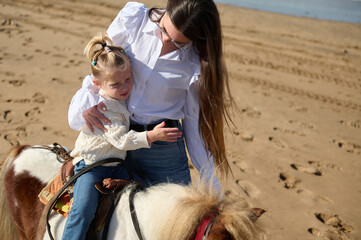 Little child girl feeling worried and hugging her mother while riding pony on the sandy beach