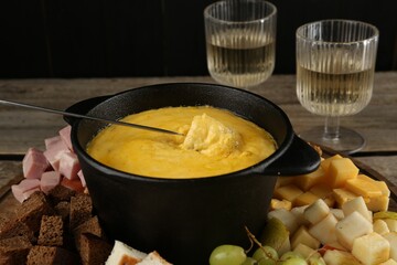 Fondue pot with melted cheese, glasses of wine and different products on wooden table, closeup