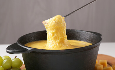 Dipping piece of bread into fondue pot with tasty melted cheese at table against gray background,...