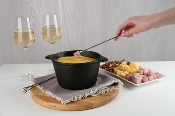 Dipping piece of ham into fondue pot with tasty melted cheese at white table, closeup