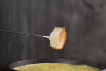 Dipping piece of bread into fondue pot with tasty melted cheese against dark gray background,...