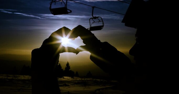 Silhouette of a girls hands creatively framing the sun within a hearth shape. Ski lift chairs and a snowy landscape in the background during golden hour. Recorded at 120fps cinema camera.