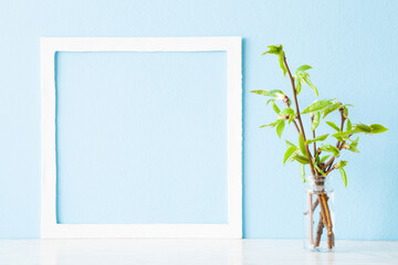 Small twigs of fresh green leaves in glass vase on wooden table at light blue wall background. Pastel color. Spring time. Empty place for inspirational text in white frame. Front view. Closeup.