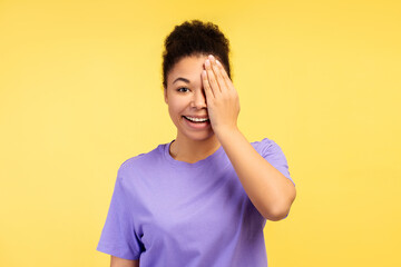 Youthful African American woman, playfully hiding one eye with her hand, on bright yellow background