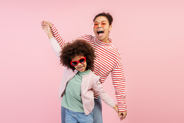 Charming mom and her young daughter in sunglasses and playfully posing isolated on pink background