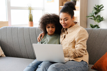 African American mom and her little girl, sitting closely on couch with laptop